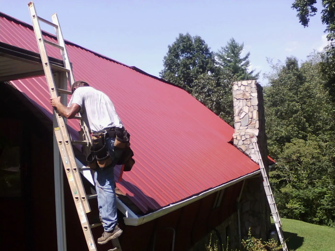 Roofer on a ladder beside a red metal roof and stone chimney — Soffit & Fascia Repair by Roofing Brothers in Pardeeville